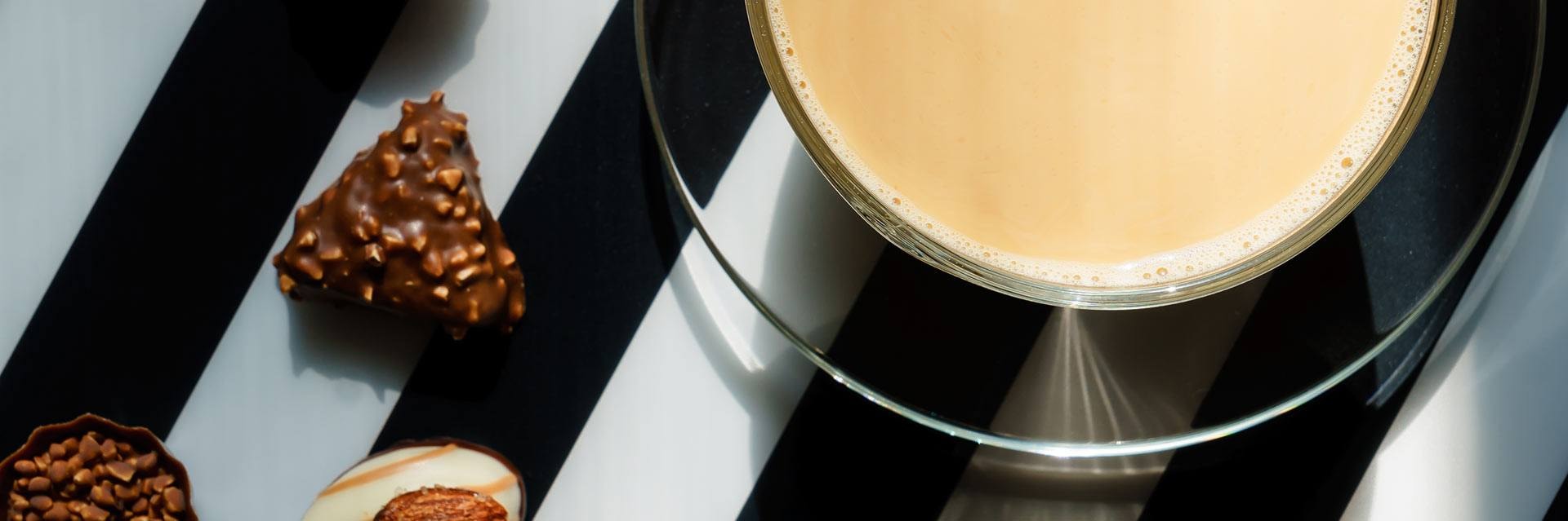 Vue de dessus d'une tasse en verre transparent contenant du café chaud au lait et des bonbons au chocolat, sur fond noir et blanc.